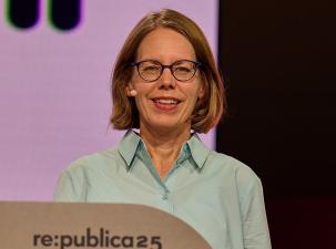 Anne Brorhilker auf der Republica25: Eine lächelnde Person mit Brille steht vor einem Rednerpult bei einer Veranstaltung, mit buntem Hintergrund.