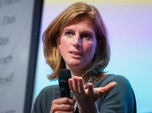 Maja Göpel auf der re:publica 19 - Frau mit blonden Haaren hält ein Mikrofon, sie gestikuliert nachdenklich während einer Diskussion. Bunte Hintergrundbeleuchtung.