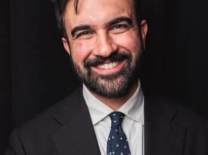 Zohran Mamdani trägt einen schwarzen Anzug und ein weißes Hemd und lächelt vor einem schwarzen Hintergrund freundlich in die Kamera.