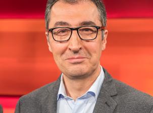 Cem Özdemir in 2020 - Ein Mann mit Brille und einem grau-melierten Anzug, lächelt vor einem orangefarbenen Hintergrund.