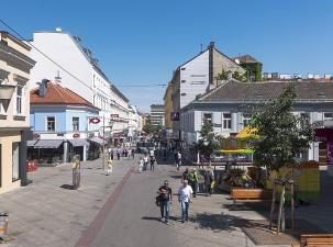 Belebte Straße mit Geschäften, Cafés und Spaziergängern unter klarem blauen Himmel.