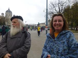 Rosi Krenn mit Matthias Reichl in Wien 2014 - Ein älterer Mann mit langem weißen Bart und eine Frau mit lockigem Haar in einem blauen Pullover stehen in einem Stadtpark, umgeben von Menschen.