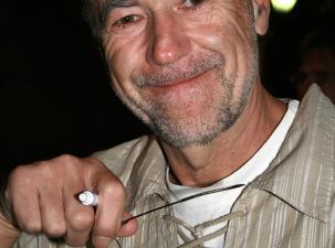 Der österreichischen Musiker Wilhelm 'Willi' Resetarits (Wien 2007) - Ein lächelnder Mann hält eine Brille in der Hand. Er trägt ein lockeres, gestreiftes Hemd und hat einen grauen Bart. Hintergrund ist unscharf.